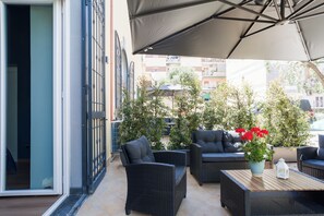 Un espace extérieur aménagé avec des meubles en osier, un pot de fleurs orné de fleurs rouges et un grand parasol offrant de l’ombre.