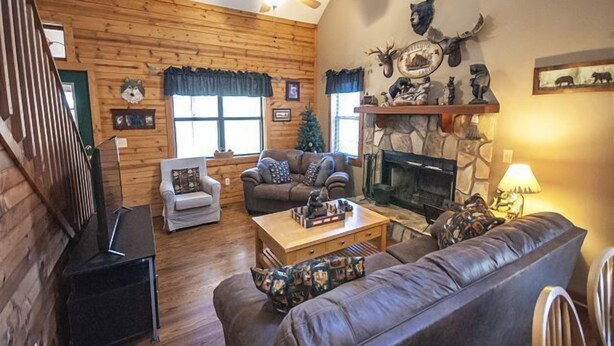A cozy log cabin interior with a fireplace, wooden walls, a ceiling fan, and a Christmas tree.