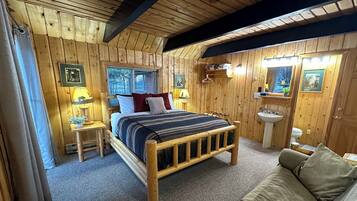 Un dormitorio de cabaña de madera con cama, mesitas de noche, un espejo y un baño con lavabo e inodoro.