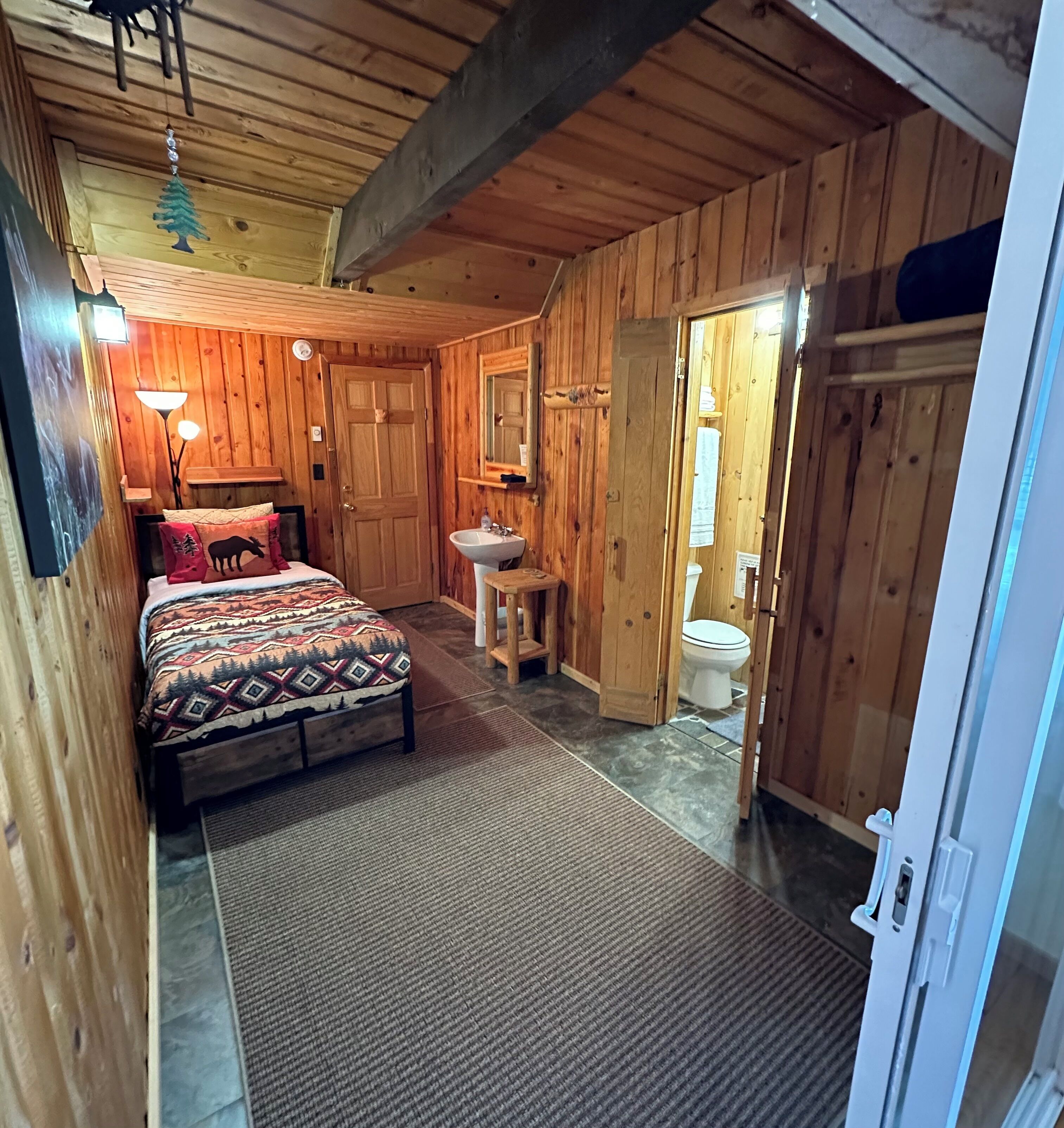 A wooden cabin bedroom with a bed, a nightstand, a lamp, a mirror, and a door leading to a bathroom.