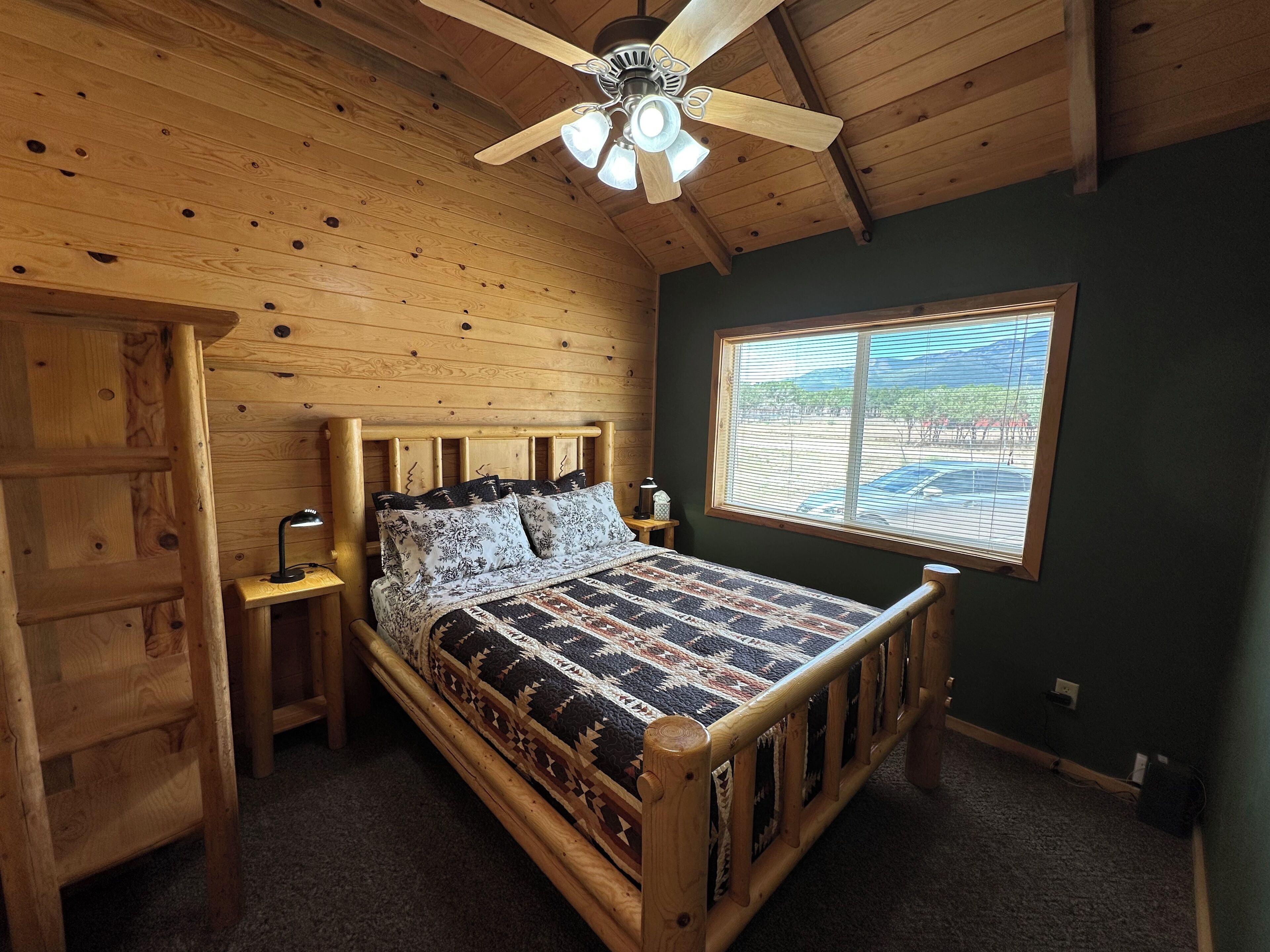 A wooden cabin bedroom with a bed, a nightstand, a window with a view, and a ceiling fan.