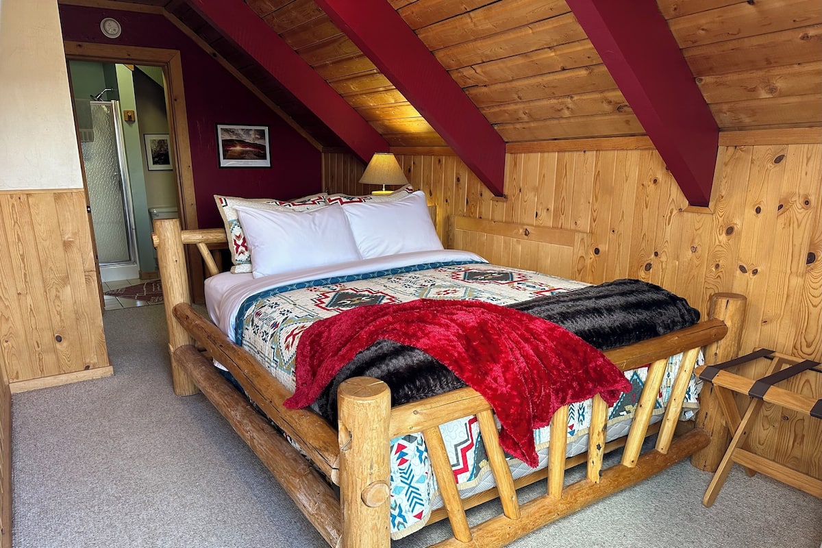 A wooden bed with a patterned bedspread and pillows, a red throw blanket, and a wooden headboard.