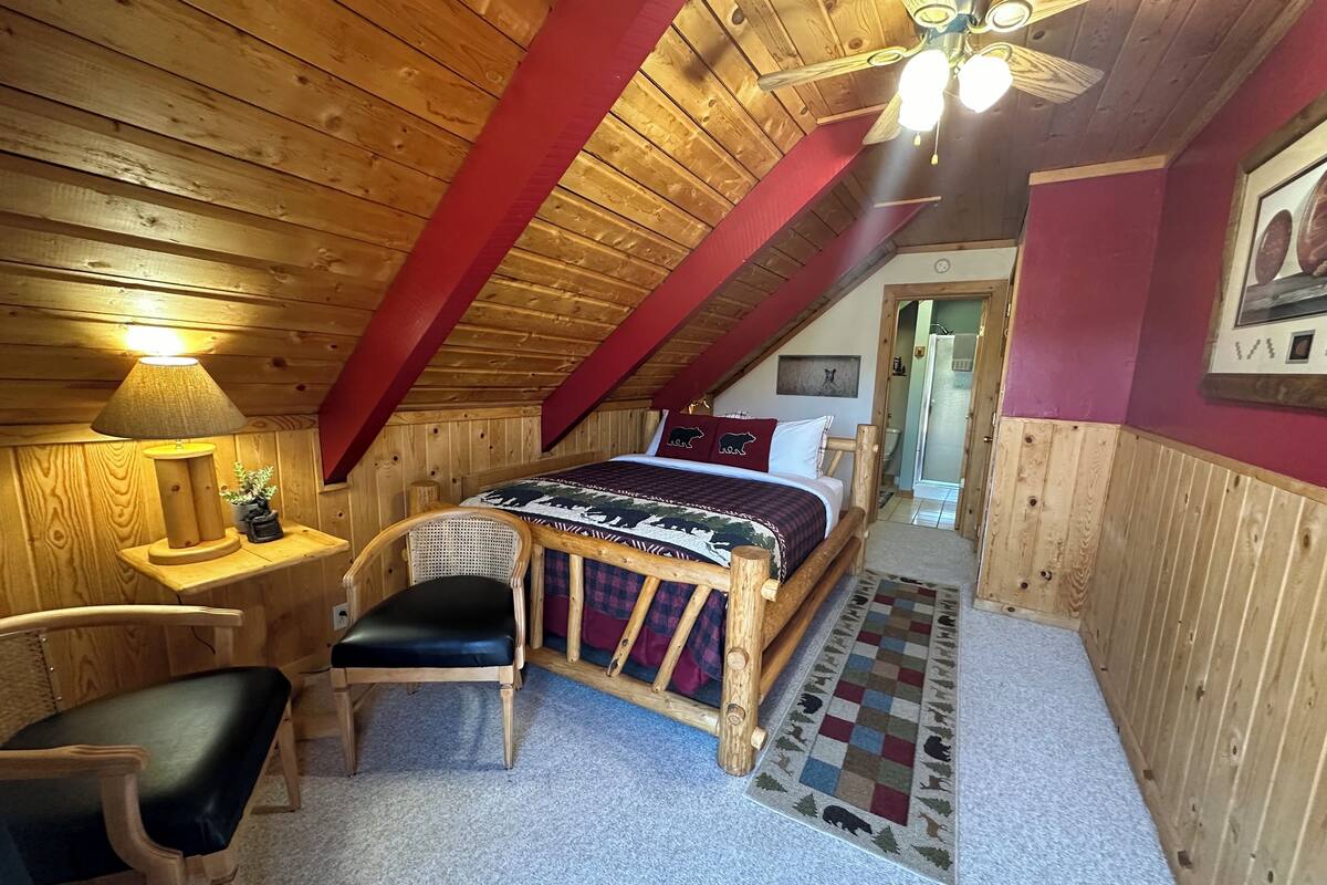 A cozy attic bedroom with a wooden ceiling, a bed with a patterned bedspread, a chair, a nightstand with a lamp, and a small potted plant.
