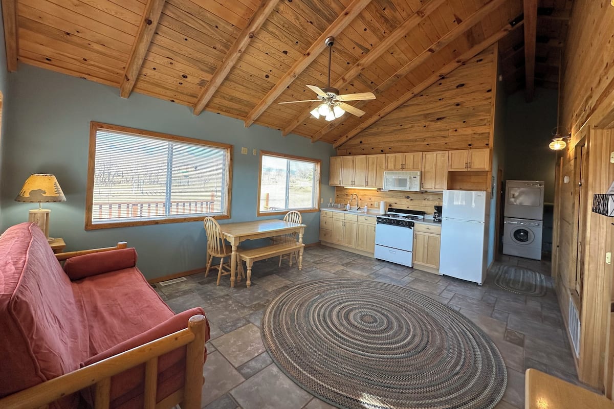 A cozy kitchen with wooden cabinets, a dining area with a table and chairs, a ceiling fan, and a large window with blinds.