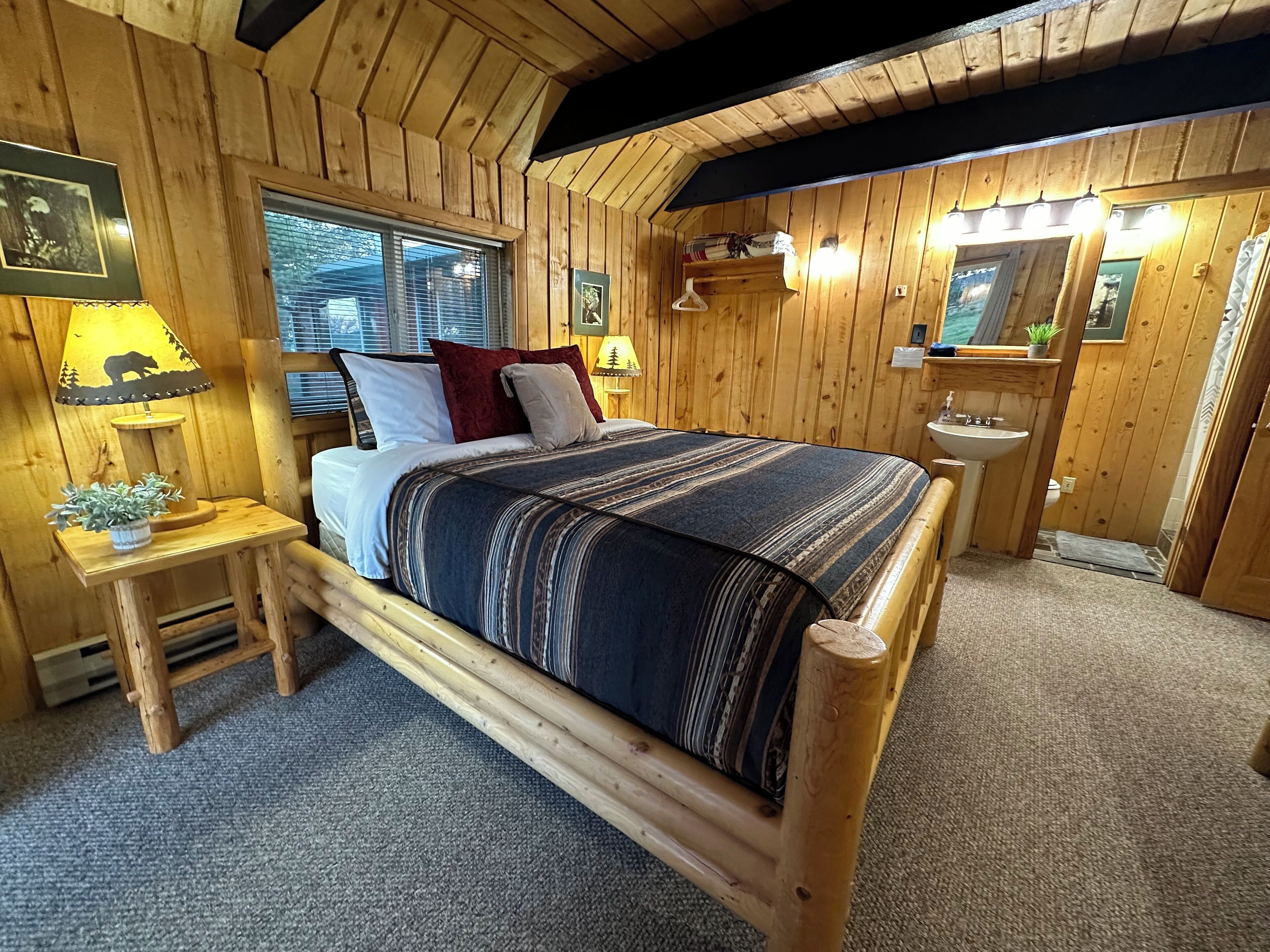 Een slaapkamer in een houten hut, met een bed, nachtlampjes, een raam en een klein wastafeltje.