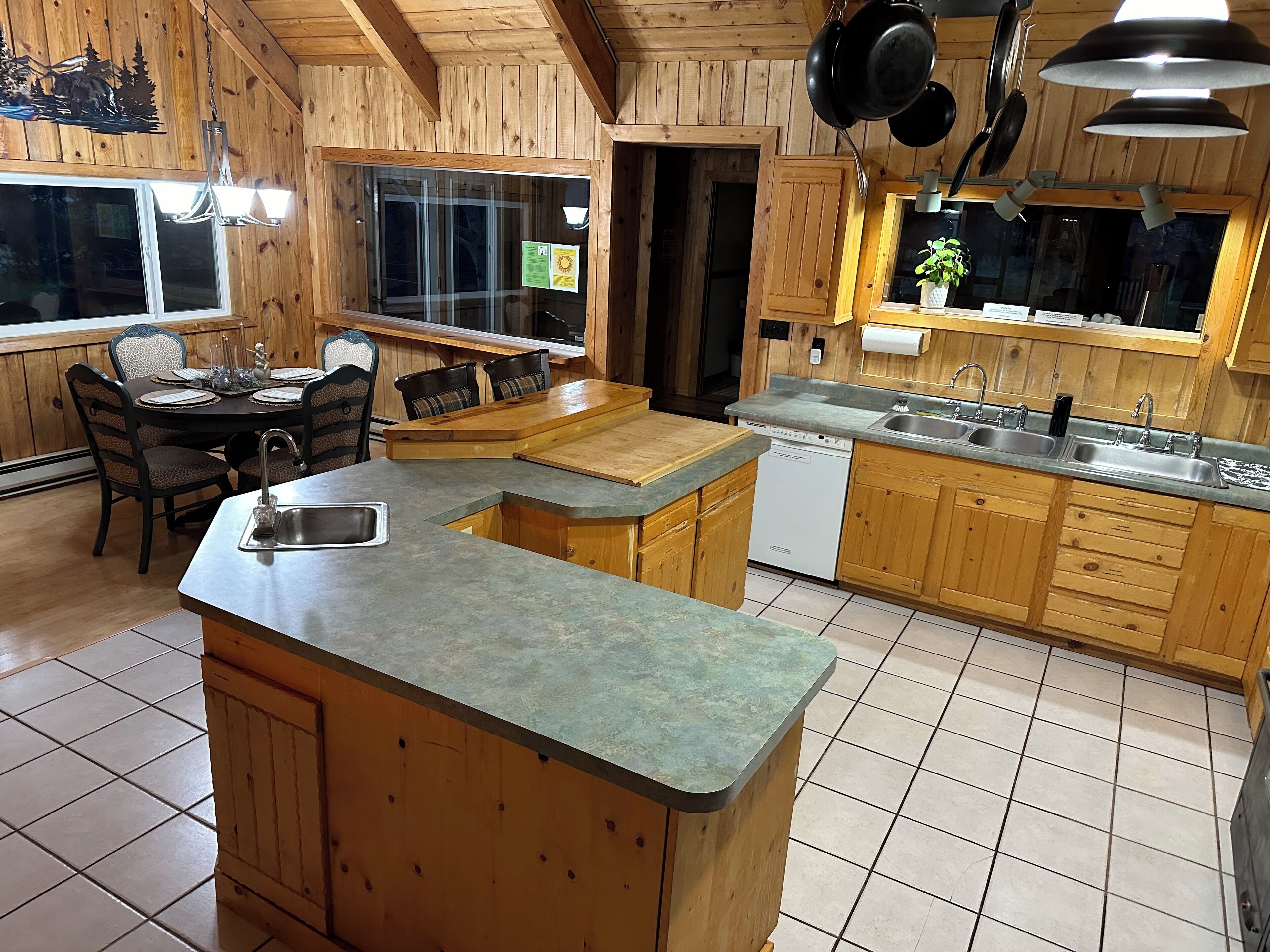 A wooden cabin kitchen with a central island, a dining area with chairs, and a large window.
