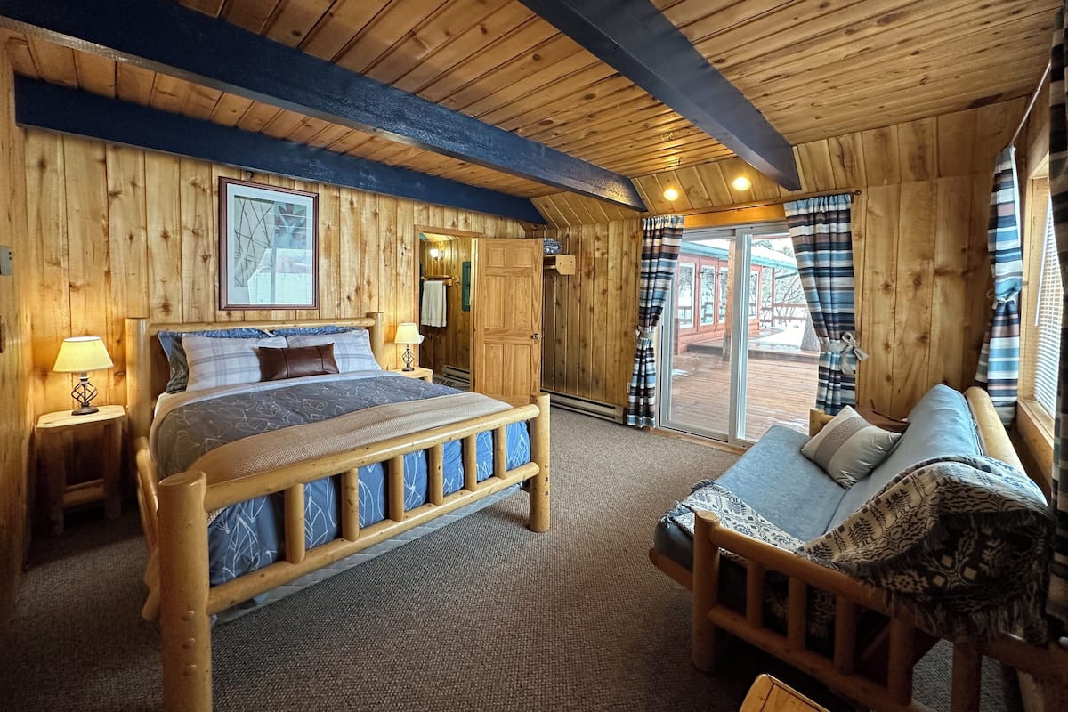 A wooden cabin bedroom with a bed, a sofa, a nightstand, and a door leading to an outdoor area.