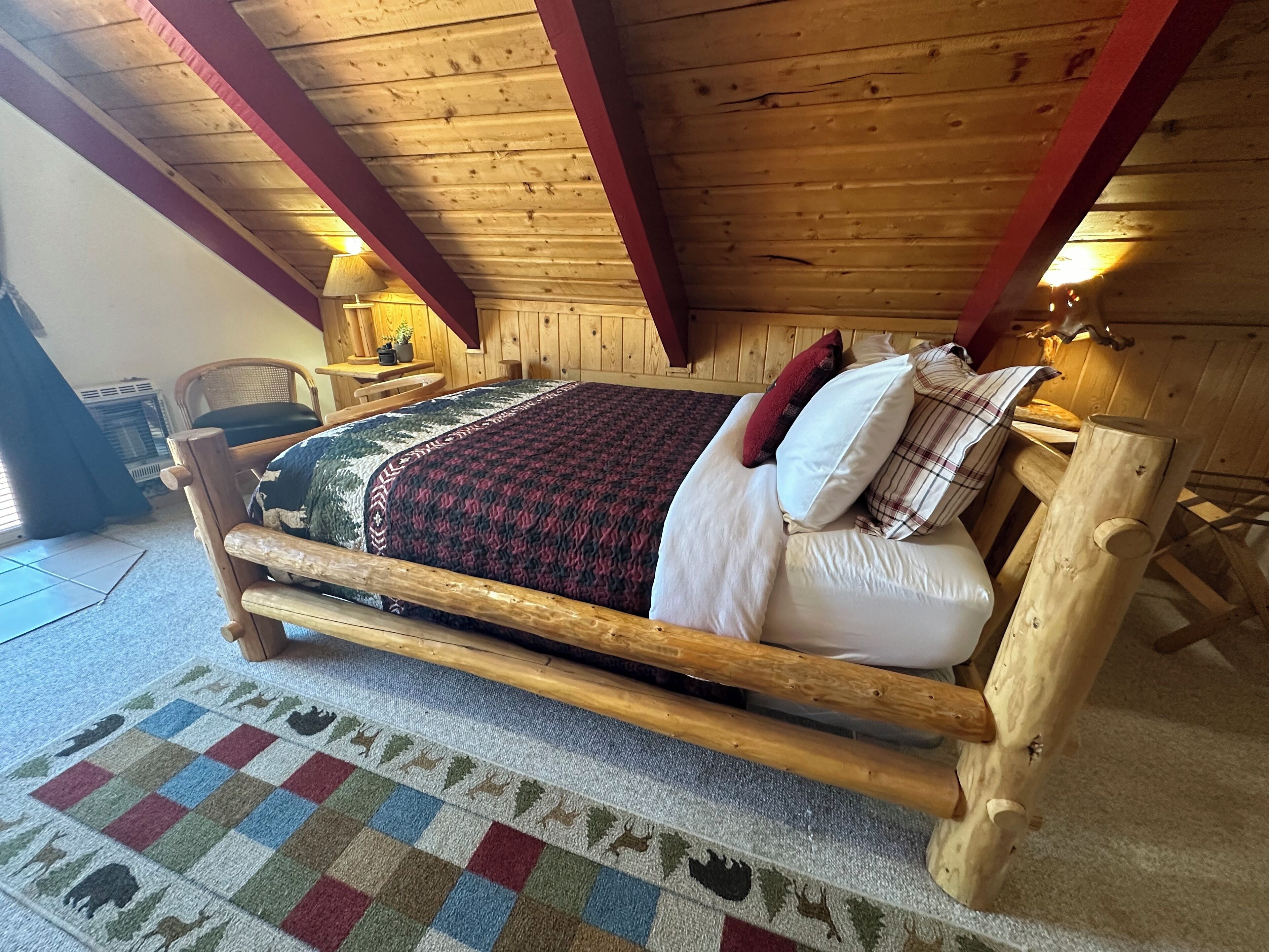 A wooden bed with a checkered rug, a lamp, and a chair in a room with wooden walls and ceiling beams.