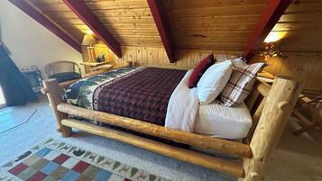 A wooden bed with a checkered rug, a lamp, and a chair in a room with wooden walls and ceiling beams.