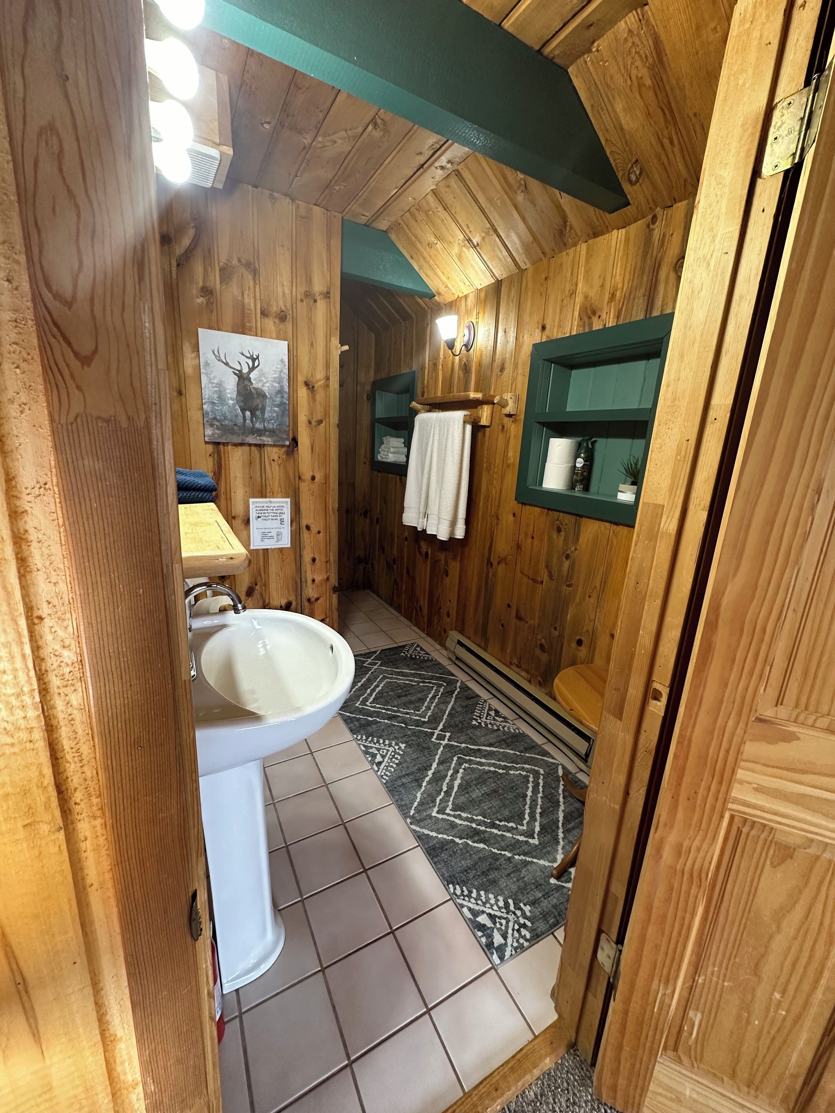 Bagno di una baita in legno con lavandino, porta asciugamani e un quadro di un cervo appeso al muro.