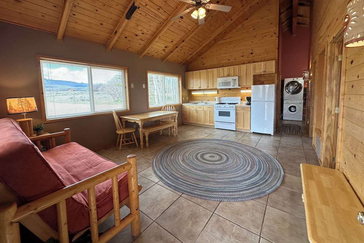 A cozy cabin interior with a dining area, kitchen, and a view through large windows.