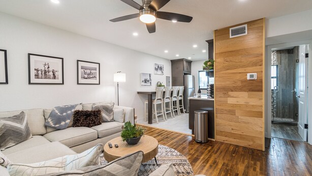 Una sala moderna con sofá, mesa de centro y ventilador de techo. Se observan cuadros enmarcados en la pared y una zona de cocina al fondo.