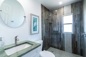 A bathroom with a round mirror, a framed artwork, a sink with a marble countertop, a toilet, and a shower with a glass door. - 400 South (Cocoa Beach)