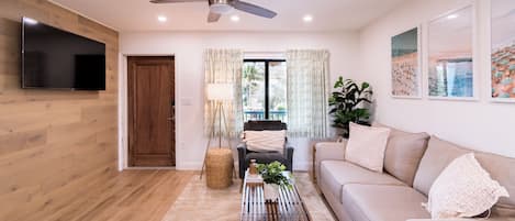 A living room with a flat-screen TV, ceiling fan, wooden flooring, beige sofa, and a coffee table with decorative items.