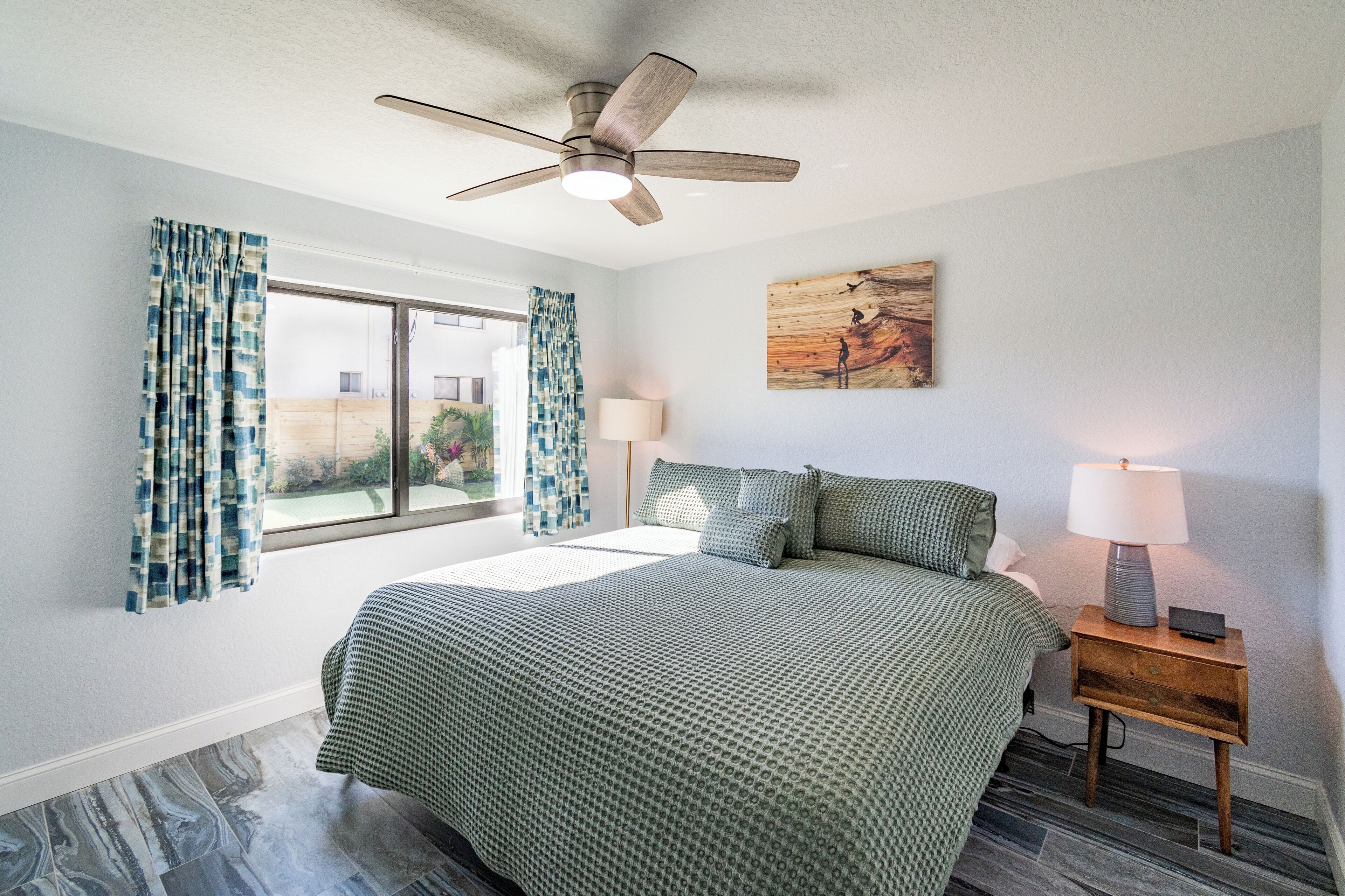 A bedroom with a bed, a ceiling fan, a window with curtains, a bedside table with a lamp, and a painting on the wall.