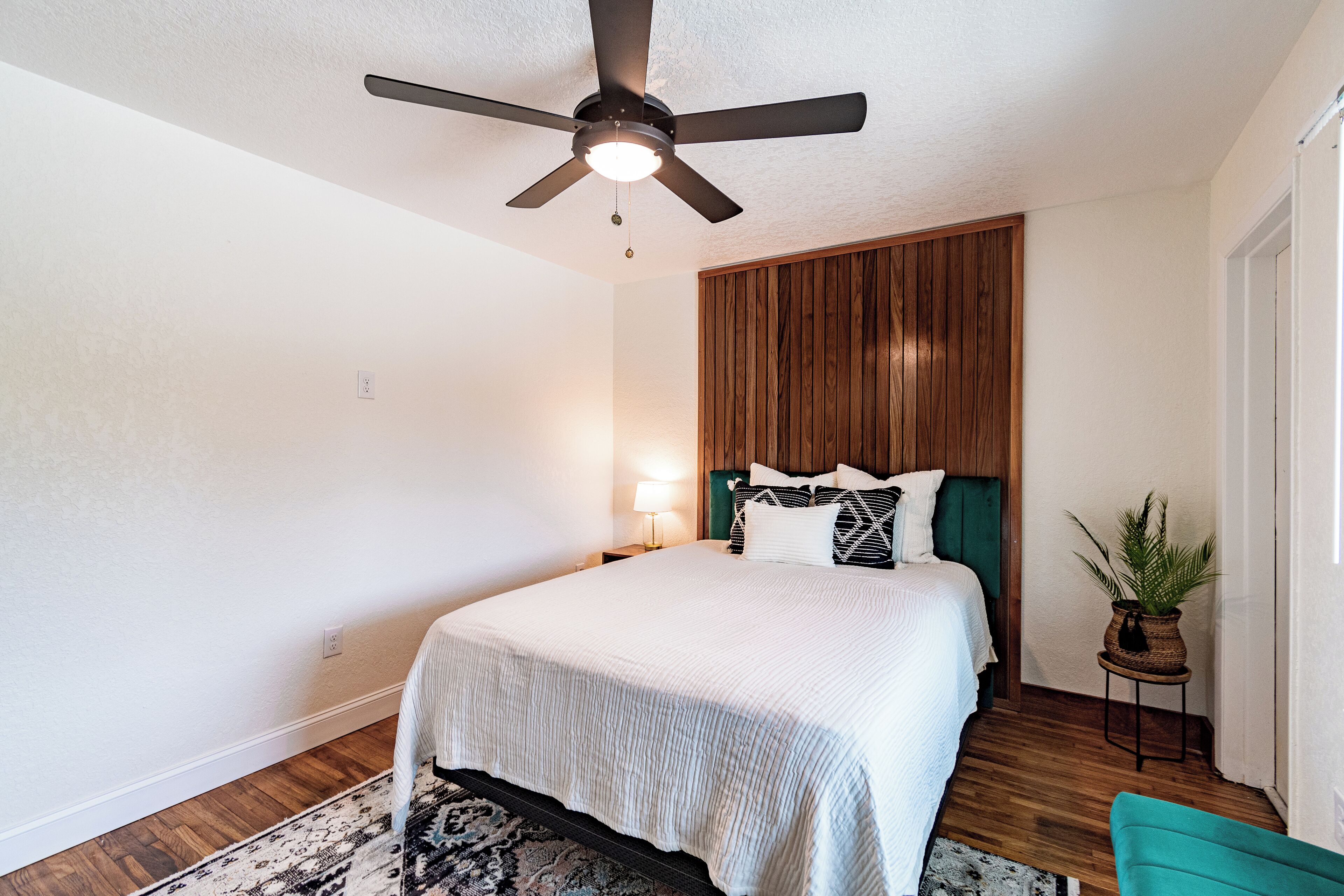 Une chambre à coucher avec un lit, un ventilateur de plafond, une table de chevet, une lampe et une plante en pot.