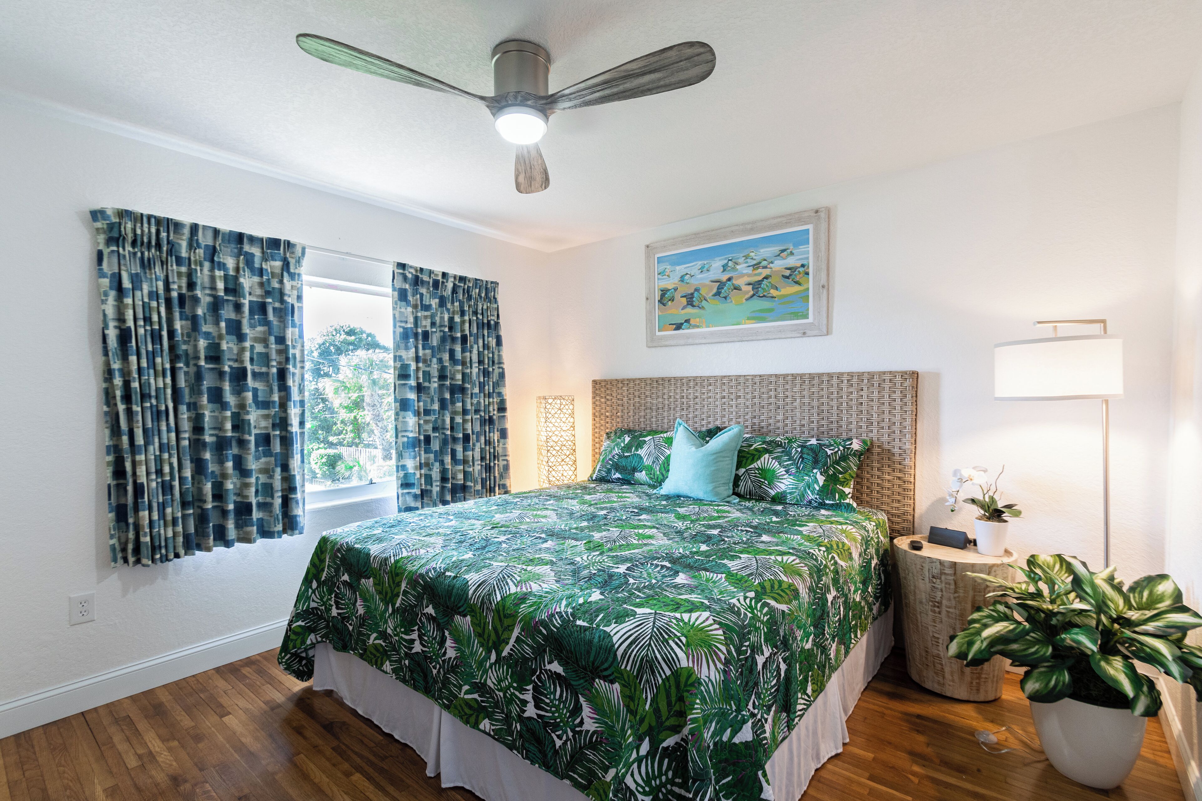 A bedroom with a bed, curtains, a ceiling fan, a nightstand, a lamp, and a potted plant.