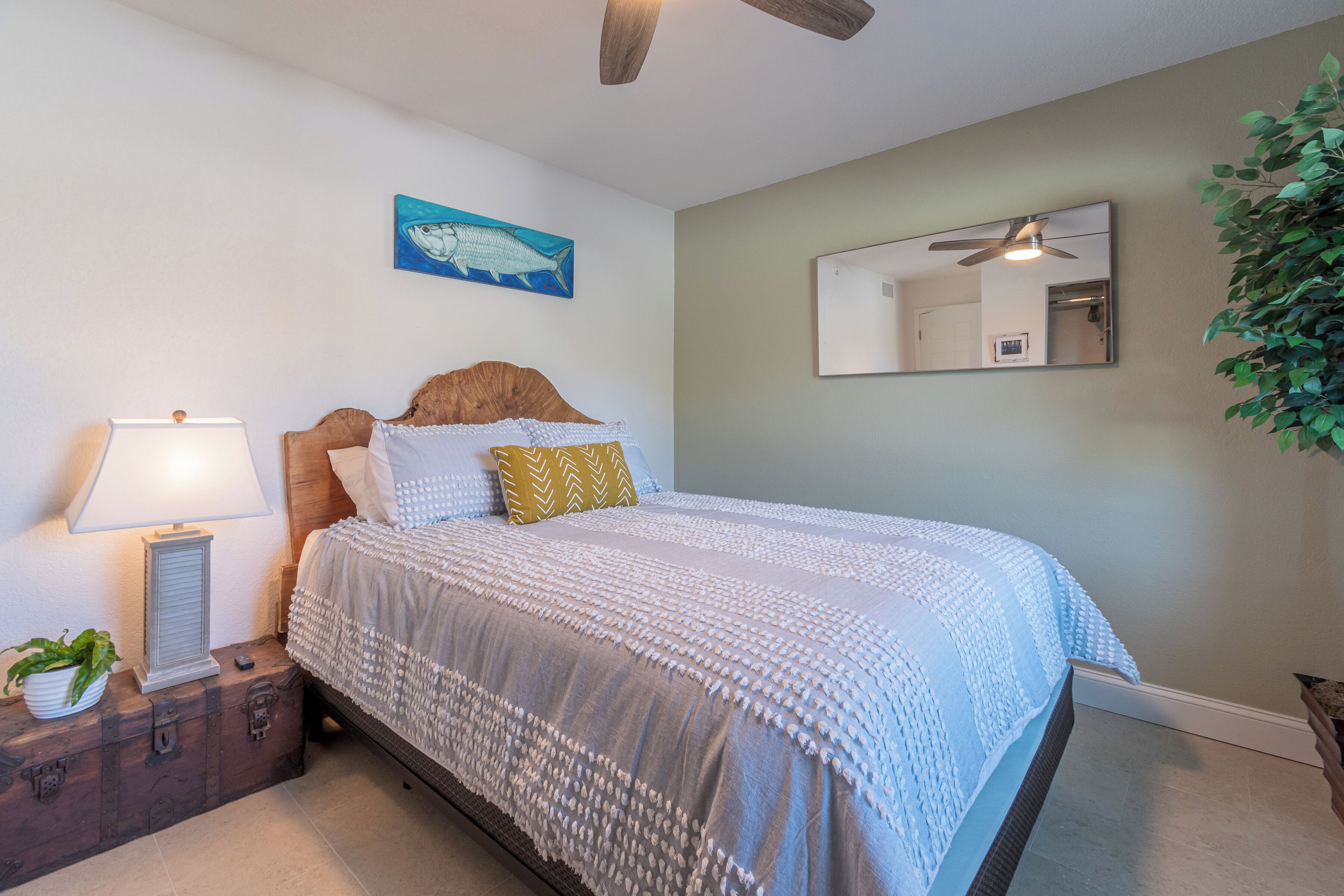 A neatly arranged bedroom with a bed, a nightstand with a lamp, a wall-mounted mirror, and a potted plant.