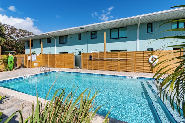A swimming pool with a wooden fence, a building with multiple windows, and a clear blue sky. - 400 South (Cocoa Beach)