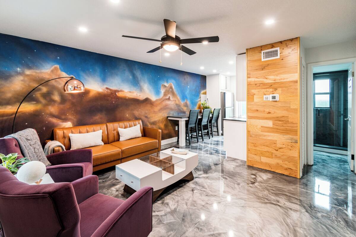 Un salon moderne avec une fresque murale représentant des étoiles, un canapé en cuir marron, des fauteuils violets et une table basse en verre.