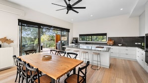 A modern kitchen with a central island, large windows, and a dining area with a wooden table and chairs. - Coastal View (Lorne)