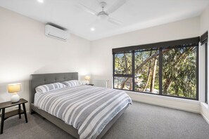 Un dormitorio con una cama, dos mesitas de noche, un ventilador de techo y un ventanal con vistas a árboles.