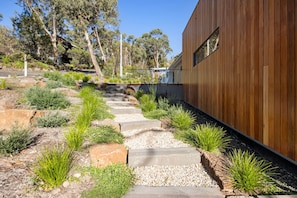 Ein modernes Gebäude mit Holzfassade, einem Betonweg zum Eingang und einem angelegten Garten mit verschiedenen Pflanzen und Steinen.