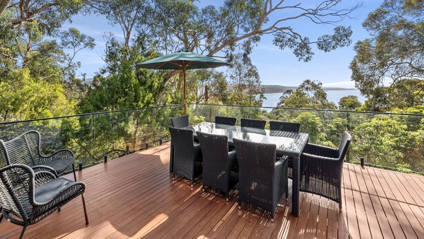 A wooden deck with a glass table and chairs, overlooking a body of water and trees. - Coastal View (Lorne)