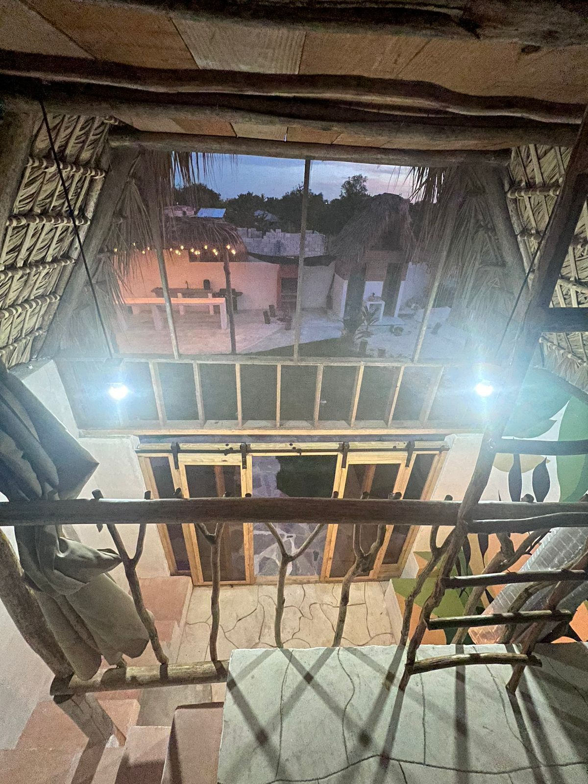 A room with a view of an outdoor area, a window with a metal frame, and a thatched roof structure outside.