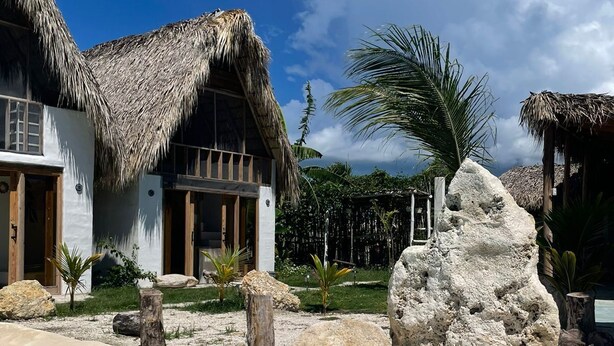 Una propiedad frente a la playa con edificios de techo de paja, una piscina cristalina y un paisaje rocoso.