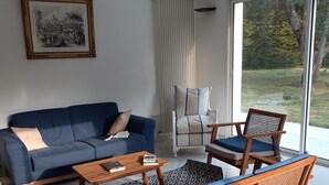 Un salon moderne avec un canapé bleu, une table basse en bois et des fauteuils en rotin. Un tableau encadré est accroché au mur, et une fenêtre offre une vue sur la verdure à l’extérieur.