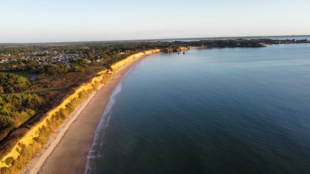 Vue aérienne d’une zone côtière avec une plage de sable, une route qui longe le rivage, et une vue lointaine d’une ville ou d’une agglomération de l’autre côté de l’eau.