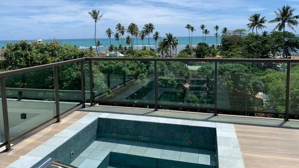 A rooftop pool with glass railings, surrounded by lush greenery and palm trees, overlooking a beach. - Flat 2 quartos no Dubourcq Residence  (Ipojuca)