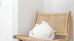 Une chaise en osier avec un coussin blanc, placée contre un mur blanc et au-dessus d’un plancher en bois.