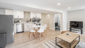 Une cuisine moderne avec des meubles blancs, un coin repas doté d’une table ronde et de chaises blanches, et un téléviseur dans le salon.