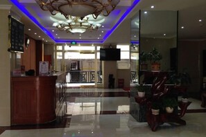Un hall de hotel con mostrador de recepción, candelabro, separador de vidrio y plantas en macetas.