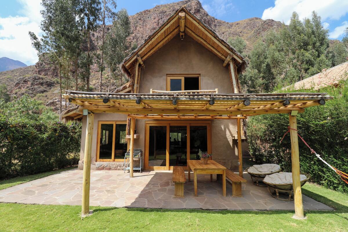 A rustic mountain lodge with a stone patio, wooden furniture, and a thatched roof.