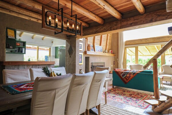 Un intérieur rustique avec des poutres en bois, un coin repas avec des chaises blanches, un tapis coloré et une vue sur l’extérieur.