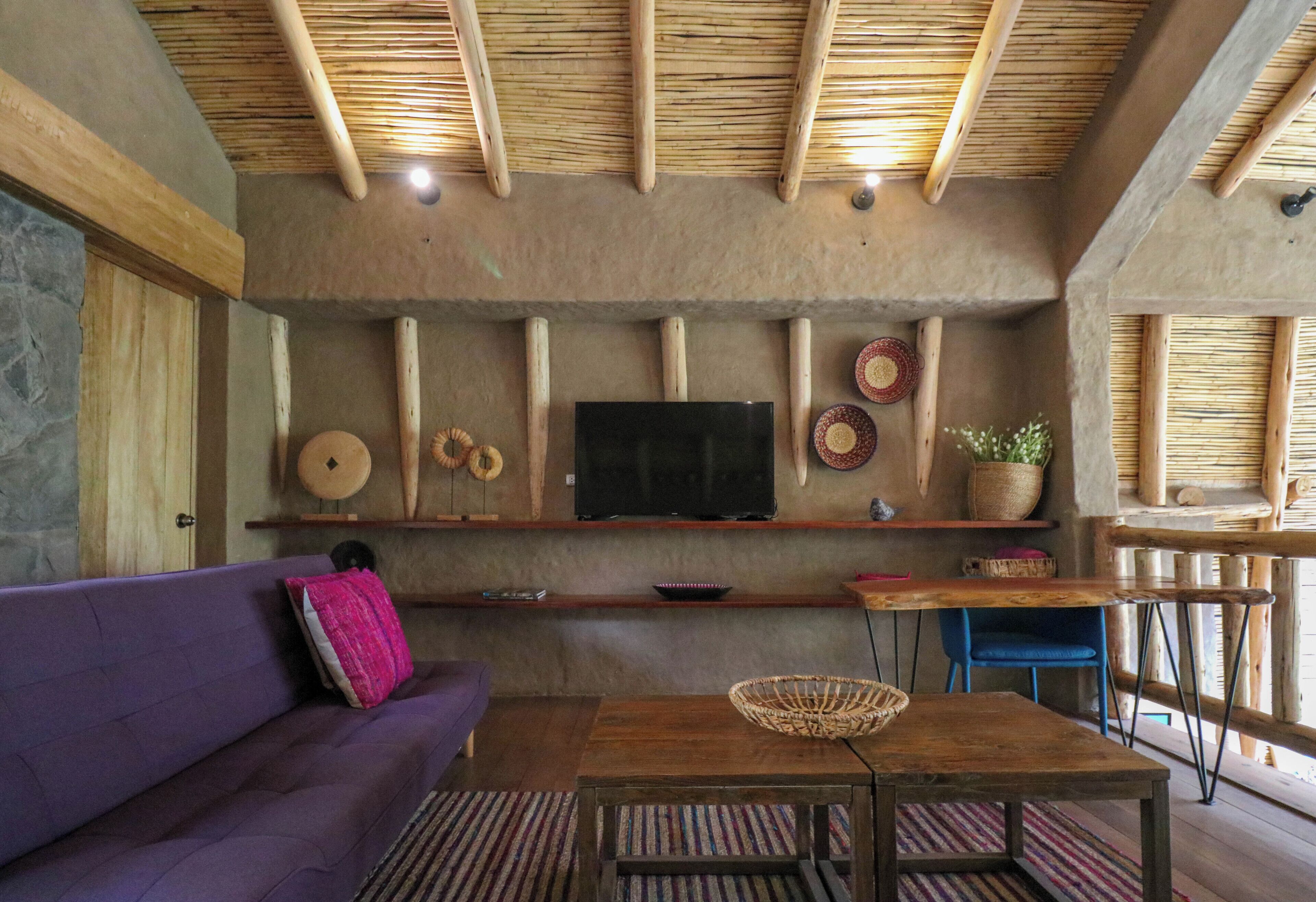 Ein rustikales Interieur mit Steinwand, Holzbalken, einem Flachbildfernseher, einem violetten Sofa und einem Holztisch mit einem gewebten Korb.