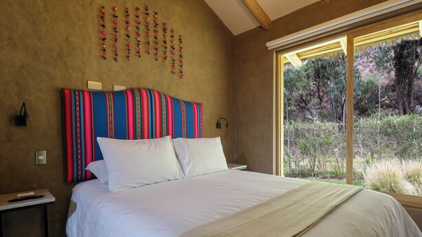 A bedroom with a large bed, a headboard with colorful stripes, a bedside table, and a window with a view of trees and grass. - Mayupata in Calca by Wynwood House (Calca)