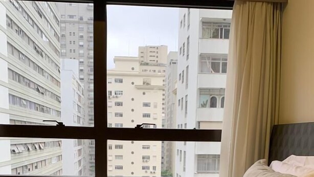 Habitación de hotel con un amplio ventanal, una cama con ropa de cama color beige y vistas a edificios altos.