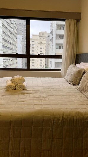 Habitación de hotel con un amplio ventanal, una cama con ropa de cama color beige y vistas a edificios altos.