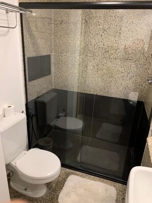 Un baño moderno con cabina de ducha de vidrio, inodoro blanco y una alfombra pequeña blanca en el suelo.