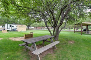 Une table de pique-nique sous un arbre, dans un espace verdoyant, avec un coin salon abrité et une remorque en arrière-plan.