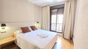 A bedroom with a bed, two pillows, a nightstand with a lamp, a window with curtains, and a wooden bench. - campezo (Madrid)