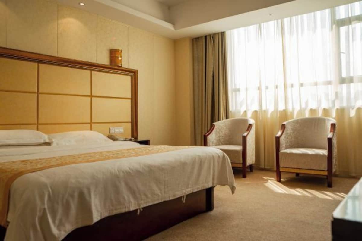 A hotel room with a large bed, two armchairs, a wooden headboard, and a large window with curtains.