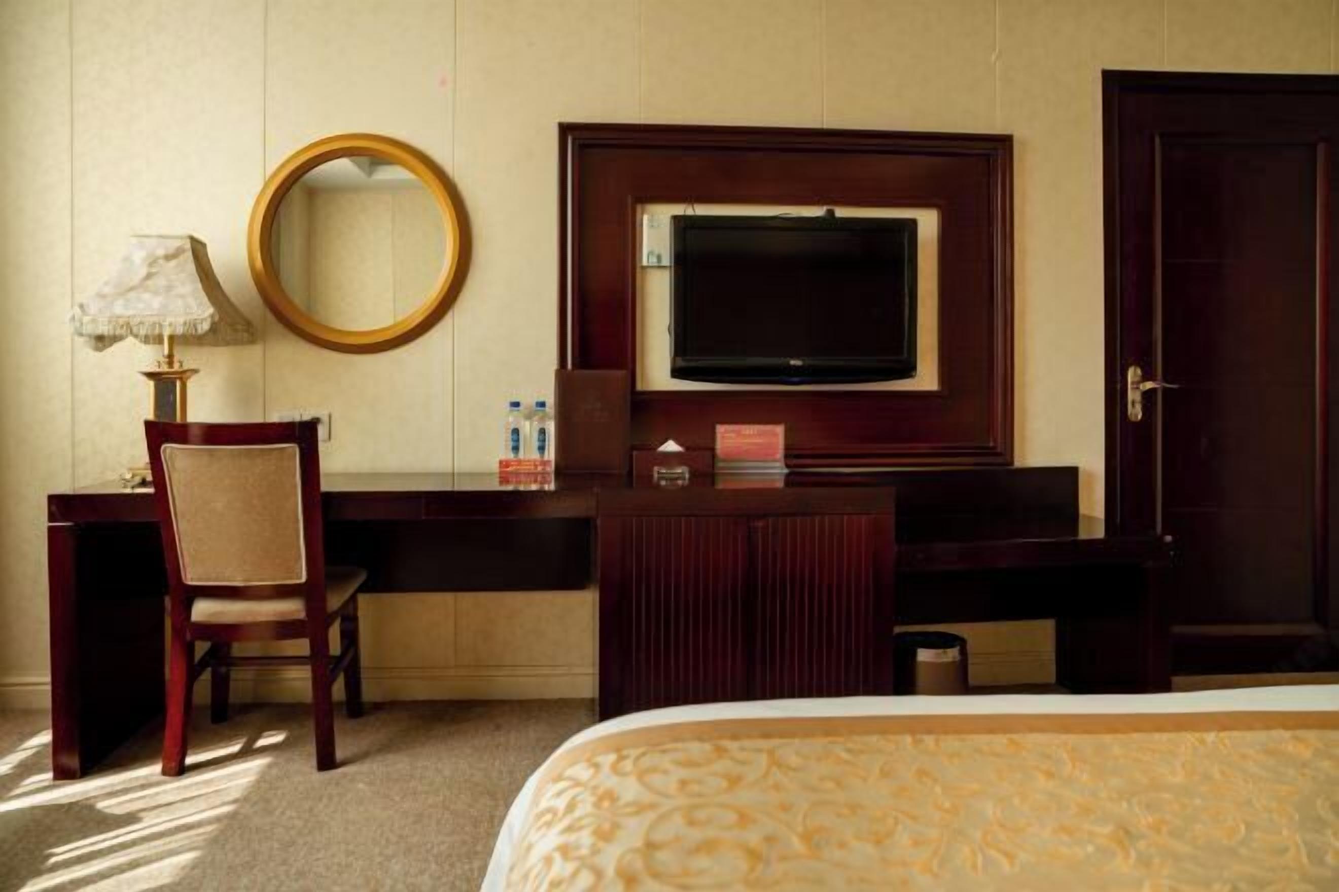 A hotel room with a bed, a desk with a chair, a television, a mirror, and a door.