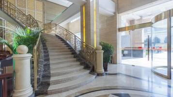 A grand staircase with marble steps and golden railings, leading to a modern lobby with a curved floor design and large glass doors.