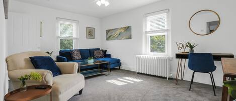 A living room with a beige sofa, a blue sofa, a coffee table, a side table with a plant, and a desk with a chair.