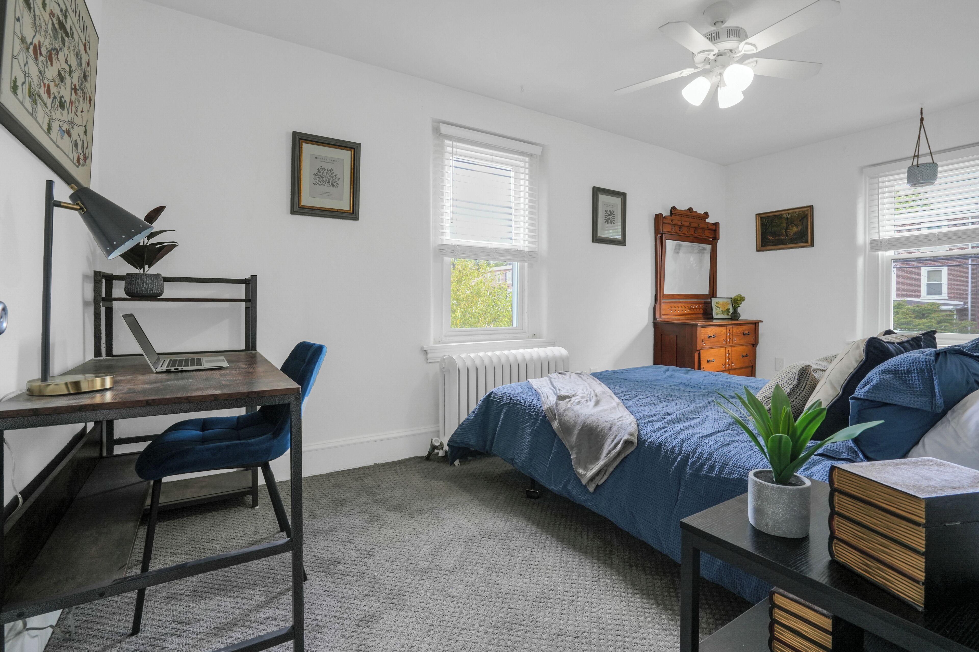 A bedroom with a bed, a desk with a chair, a radiator, a ceiling fan, and a window with blinds.
